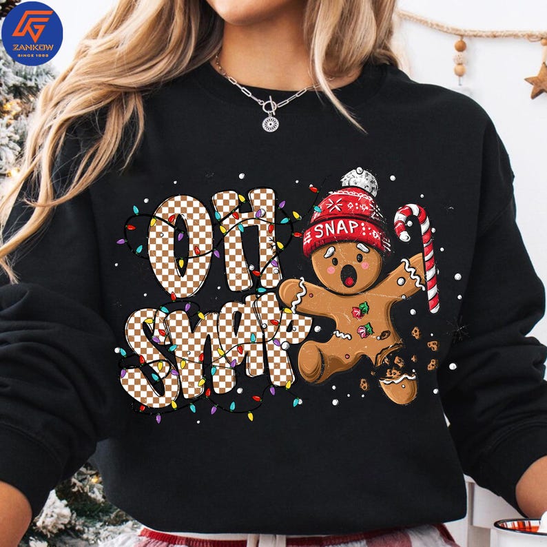 May include: Black sweatshirt with a festive design featuring the words "Oh Snap" in a checkered pattern, surrounded by colorful lights. A gingerbread man with a red hat and candy cane completes the holiday theme.