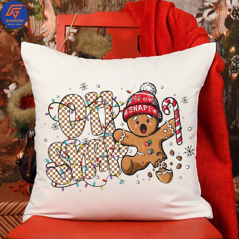 May include: White square pillow with a Christmas theme. The pillow features a gingerbread man wearing a red hat with the word "SNAP" and holding a candy cane. The words "OH SNAP" are written in a checkered pattern with Christmas lights.