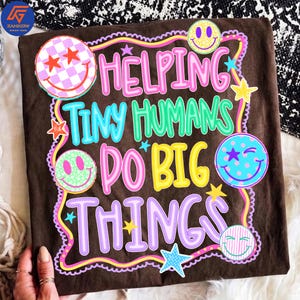 May include: A dark brown t-shirt featuring the text HELPING TINY HUMANS DO BIG THINGS in colorful, layered lettering. The design includes various smiley faces with checkered, spotted, and striped patterns, along with several small colorful stars.