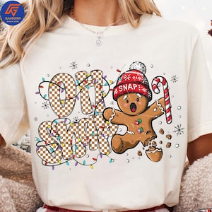 May include: Cream-colored t-shirt featuring a gingerbread man with a red knit cap and candy cane. The words "OH SNAP" are printed in a checkered pattern, surrounded by colorful Christmas lights and snowflakes.