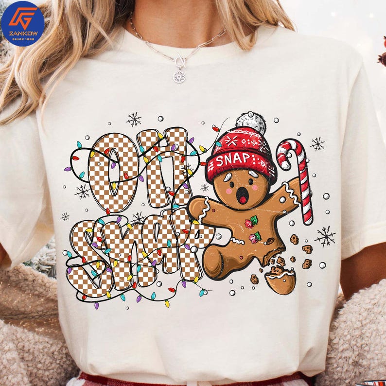 May include: Cream-colored t-shirt featuring a gingerbread man with a red knit cap and candy cane. The words "OH SNAP" are printed in a checkered pattern, surrounded by colorful Christmas lights and snowflakes.
