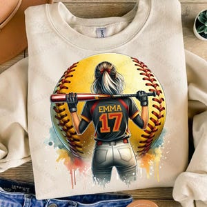K&ouml;nnte beinhalten: Ein beiger Pullover mit einem Aquarell-Design einer Softballspielerin, die einen Schl&auml;ger h&auml;lt. Die Spielerin tr&auml;gt ein schwarzes Trikot mit dem Namen "Emma" und der Nummer "17" auf dem R&uuml;cken. Der Softball ist gelb mit roten N&auml;hten.