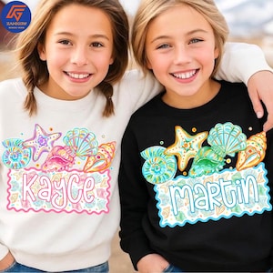 Peut inclure: Deux enfants portant des sweat-shirts assortis. L'un est blanc avec une étiquette nominative rose qui dit "Kayce" et l'autre est noir avec une étiquette nominative bleue qui dit "Martin". Les deux sweat-shirts ont un motif de coquillages et d'étoiles de mer.