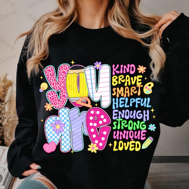 May include: Black sweatshirt with the words "You Are" in colorful, bubble letters. The words "Kind, Brave, Smart, Helpful, Enough, Strong, Unique, Loved" are listed vertically in a complementary font, with decorative stars and flowers.
