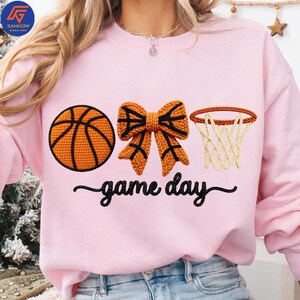 May include: A light pink sweatshirt with a basketball, bow, and hoop design. The basketball is orange with black lines, the bow is orange with black accents, and the hoop is brown with a white net. The words "game day" are written in black script.