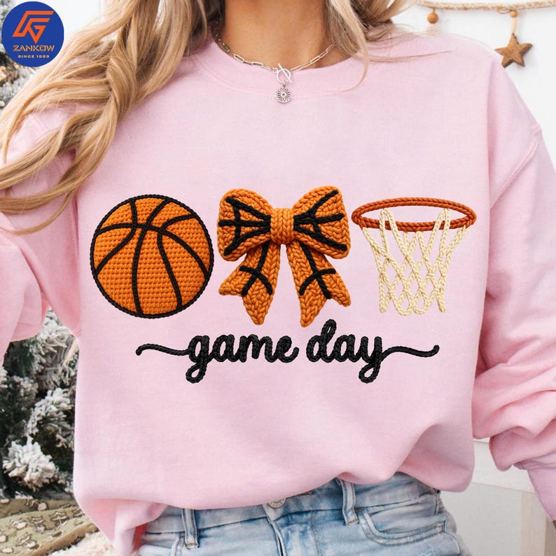May include: A light pink sweatshirt with a basketball, bow, and hoop design. The basketball is orange with black lines, the bow is orange with black accents, and the hoop is brown with a white net. The words "game day" are written in black script.