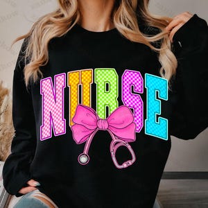 K&ouml;nnte beinhalten: Schwarzes Sweatshirt mit dem Wort "NURSE" in bunten, karierten Buchstaben. Eine rosa Schleife und eine Stethoskop-Grafik sind unter dem Wort zentriert. Das Sweatshirt ist ein Rundhals-Stil.