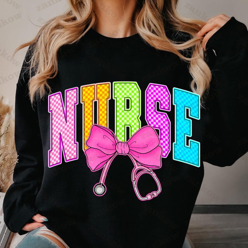 K&ouml;nnte beinhalten: Schwarzes Sweatshirt mit dem Wort "NURSE" in bunten, karierten Buchstaben. Eine rosa Schleife und eine Stethoskop-Grafik sind unter dem Wort zentriert. Das Sweatshirt ist ein Rundhals-Stil.