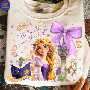May include: Cream-colored sweatshirt featuring a graphic of a princess with long blonde hair, wearing a purple dress, and the text "At last I see the light." The design includes a castle, horse, and other fairytale elements.