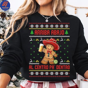 May include: Black sweatshirt with a festive design featuring a gingerbread man wearing a red sombrero and holding a beer. The text "ARRIBA ABAJO" and "AL CENTRO PA' DENTRO" are displayed above and below the gingerbread man, with decorative elements.