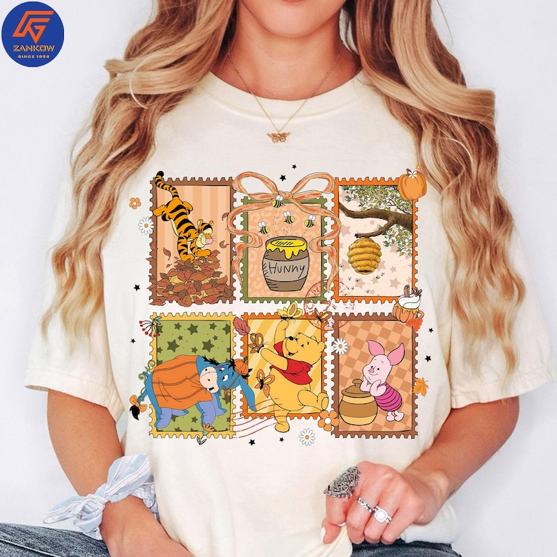 May include: Cream-coloured t-shirt featuring a colourful graphic of Winnie the Pooh characters in postage stamp designs. The design includes Tigger, Pooh, Eeyore, Piglet, a honey pot, and a beehive. The word "Hunny" is also visible.