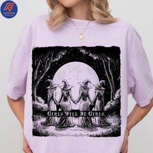 May include: Lilac-colored t-shirt featuring a black and white illustration of four witches holding hands in a forest, with a full moon in the background. The text "GIRLS WILL BE GIRLS" is printed below the image. The shirt has a crew neck and short sleeves.