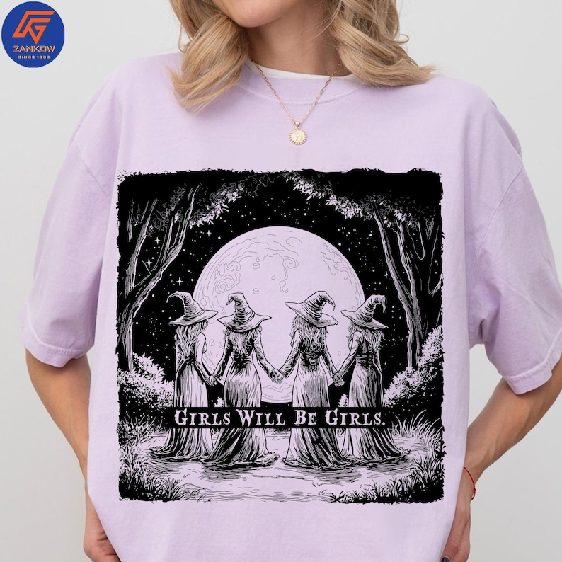 May include: Lilac-colored t-shirt featuring a black and white illustration of four witches holding hands in a forest, with a full moon in the background. The text "GIRLS WILL BE GIRLS" is printed below the image. The shirt has a crew neck and short sleeves.