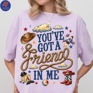 May include: A light purple t-shirt featuring a graphic print with the text "YOU'VE GOT A FRIEND IN ME" in blue letters. The design includes a brown cowboy hat, a coiled rope, a toy rocket, a block with the letter A, a ball, and cowboy boots.