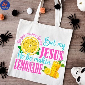 May include: White canvas tote bag with the text "Life gave me some lemons But my JESUS He be makin LEMONADE" in blue and pink. The design includes a yellow lemon slice, green leaves, and a glass of lemonade.