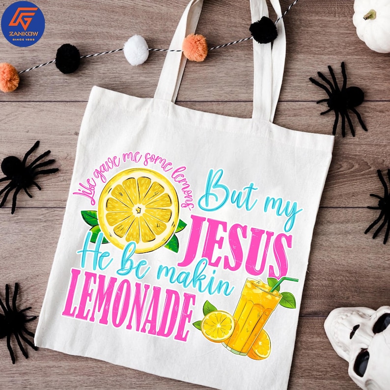May include: White canvas tote bag with the text "Life gave me some lemons But my JESUS He be makin LEMONADE" in blue and pink. The design includes a yellow lemon slice, green leaves, and a glass of lemonade.