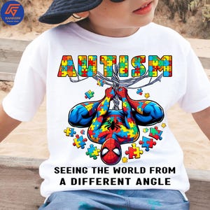 May include: White t-shirt featuring a colorful Spider-Man design made of puzzle pieces. The word "AUTISM" is above the character, also in puzzle piece style. The text "SEEING THE WORLD FROM A DIFFERENT ANGLE" is below the character.