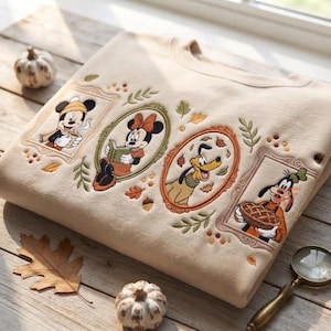 Peut inclure: Un sweat-shirt beige à col rond orné de portraits brodés de Mickey Mouse avec une tasse, Minnie Mouse lisant un livre, Pluto et Dingo tenant une tarte. Chaque personnage est encadré par une bordure ovale ou rectangulaire, entouré de feuilles d'automne.