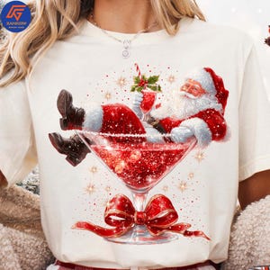 May include: Cream-colored t-shirt featuring a festive graphic of Santa Claus relaxing in a giant martini glass filled with red liquid and glitter. Santa is wearing his traditional red suit and holding a cocktail. A red bow adorns the base of the glass.