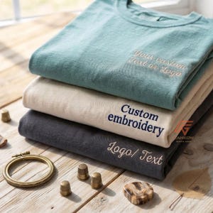 May include: A stack of three folded t-shirts in teal, beige, and charcoal grey. The teal shirt features embroidered text reading Your custom Text or Logo. The beige shirt has Custom embroidery text, and the charcoal shirt features Logo/ Text.