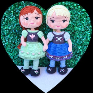 Puede incluir: Dos mu&ntilde;ecas de ganchillo hechas a mano, una con pelo rojo y un vestido verde, la otra con pelo rubio y un vestido azul, tomadas de la mano. Las mu&ntilde;ecas tienen zapatos negros y est&aacute;n colocadas sobre un fondo verde dentro de un marco en forma de coraz&oacute;n.