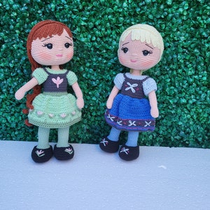 Puede incluir: Dos mu&ntilde;ecas de ganchillo hechas a mano, una con cabello casta&ntilde;o rojizo y un vestido verde, la otra con cabello rubio y un vestido azul. Ambas mu&ntilde;ecas tienen rasgos faciales detallados y llevan zapatos negros. Est&aacute;n frente a un seto verde.