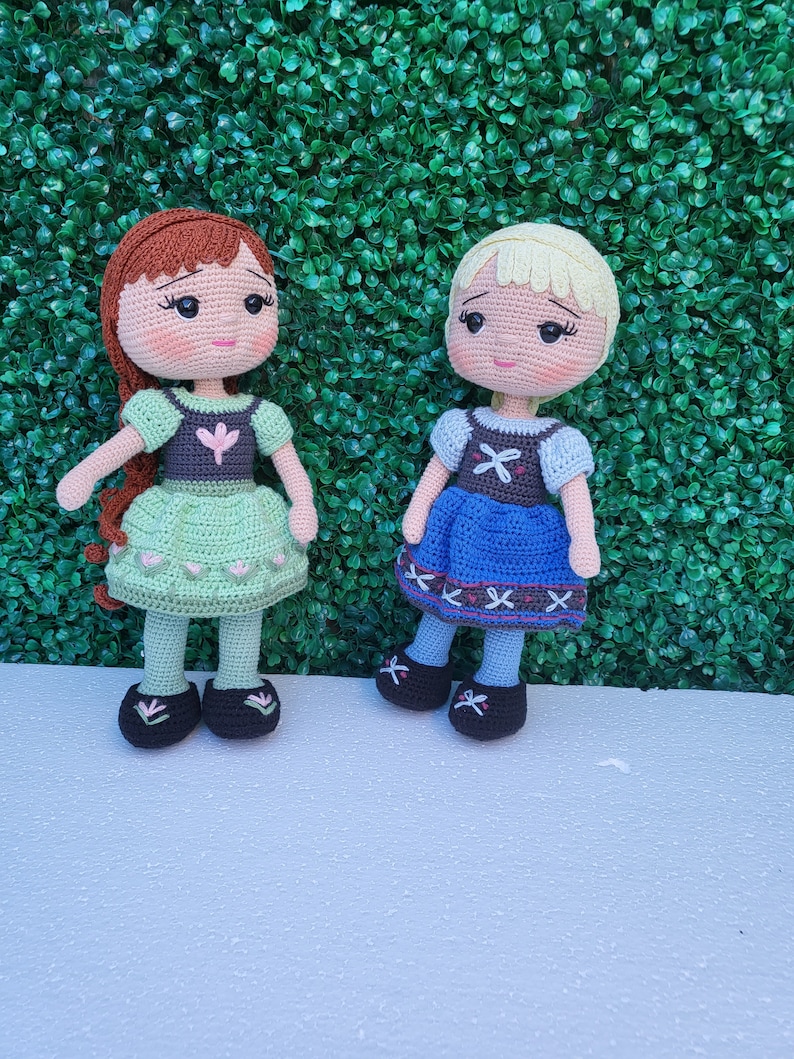 Puede incluir: Dos mu&ntilde;ecas de ganchillo hechas a mano, una con cabello casta&ntilde;o rojizo y un vestido verde, la otra con cabello rubio y un vestido azul. Ambas mu&ntilde;ecas tienen rasgos faciales detallados y llevan zapatos negros. Est&aacute;n frente a un seto verde.