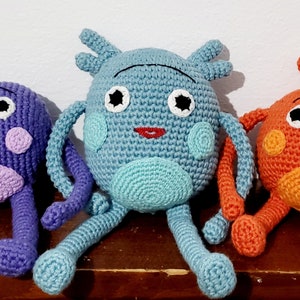 May include: Three handmade crochet dolls in purple, blue, and orange. Each doll has a round head, large eyes, and long limbs. The purple doll has a light purple belly and cheeks. The blue doll has a light blue belly and a crown. The orange doll has an orange belly.