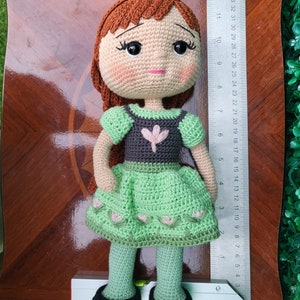 Puede incluir: Mu&ntilde;eca de ganchillo hecha a mano, cabello casta&ntilde;o y tez clara. La mu&ntilde;eca viste un vestido verde con corpi&ntilde;o gris y un adorno floral rosa. La mu&ntilde;eca mide aproximadamente 30 cm de alto, con zapatos negros y mallas verdes.