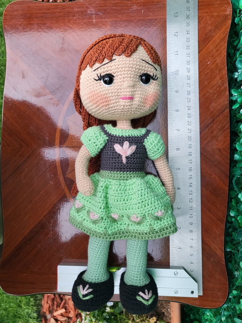 Puede incluir: Mu&ntilde;eca de ganchillo hecha a mano, cabello casta&ntilde;o y tez clara. La mu&ntilde;eca viste un vestido verde con corpi&ntilde;o gris y un adorno floral rosa. La mu&ntilde;eca mide aproximadamente 30 cm de alto, con zapatos negros y mallas verdes.