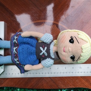 Puede incluir: Mu&ntilde;eca de ganchillo con piel clara, cabello rubio y un vestido azul. La mu&ntilde;eca tiene zapatos negros y mide aproximadamente 25 cm de largo. El vestido de la mu&ntilde;eca tiene un dise&ntilde;o decorativo. La mu&ntilde;eca est&aacute; sobre una superficie de madera.