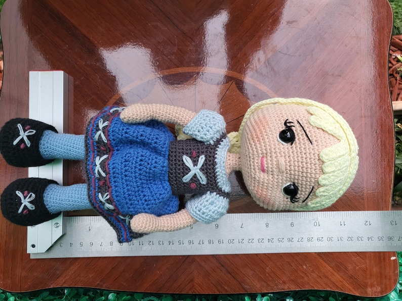 Puede incluir: Mu&ntilde;eca de ganchillo con piel clara, cabello rubio y un vestido azul. La mu&ntilde;eca tiene zapatos negros y mide aproximadamente 25 cm de largo. El vestido de la mu&ntilde;eca tiene un dise&ntilde;o decorativo. La mu&ntilde;eca est&aacute; sobre una superficie de madera.