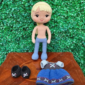 Puede incluir: Mu&ntilde;eca de ganchillo hecha a mano con cabello rubio, pantalones azules y un atuendo a juego. La mu&ntilde;eca tiene zapatos negros y se exhibe sobre una superficie de madera contra un fondo verde. El atuendo incluye un vestido azul y una parte superior gris.