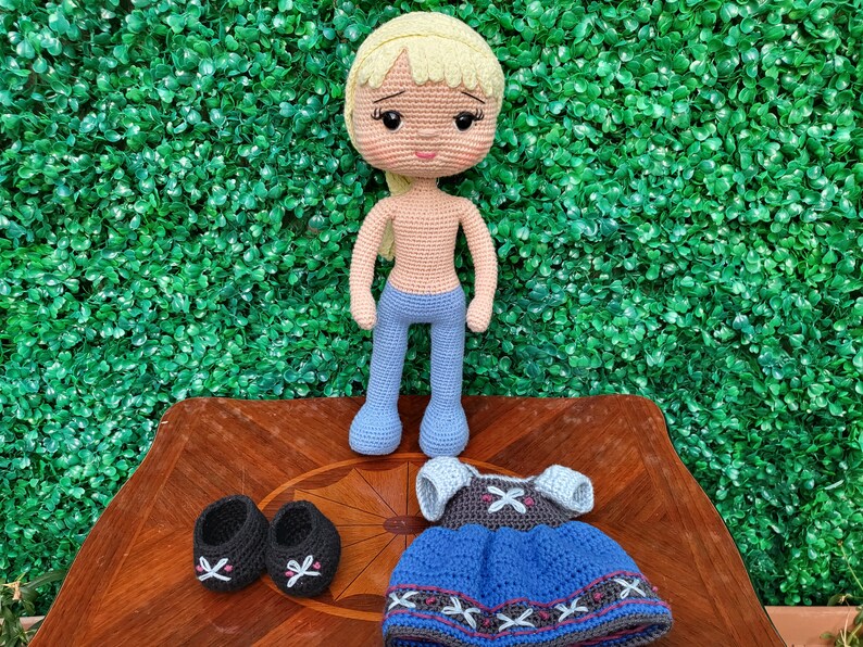 Puede incluir: Mu&ntilde;eca de ganchillo hecha a mano con cabello rubio, pantalones azules y un atuendo a juego. La mu&ntilde;eca tiene zapatos negros y se exhibe sobre una superficie de madera contra un fondo verde. El atuendo incluye un vestido azul y una parte superior gris.