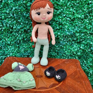Puede incluir: Mu&ntilde;eca de ganchillo hecha a mano con cabello rojo, que lleva pantalones verde claro. Los accesorios incluyen un vestido verde con detalles de flores rosas y zapatos negros con detalles florales blancos. La mu&ntilde;eca se exhibe sobre una superficie de madera.