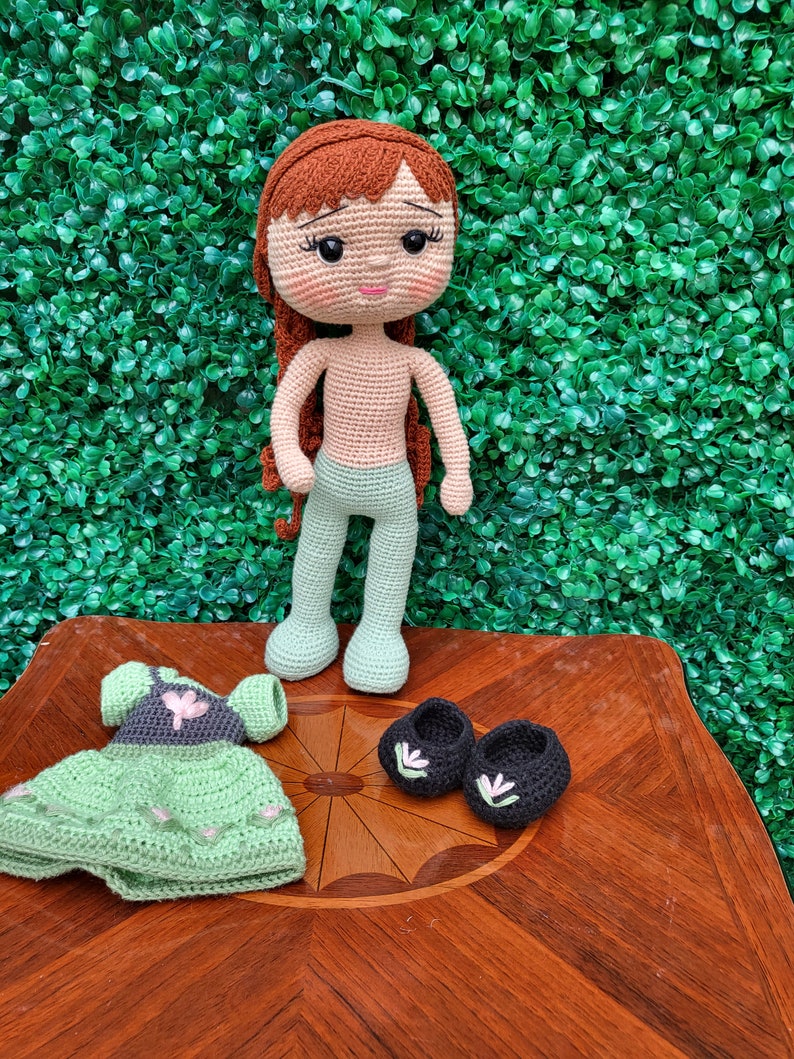 Puede incluir: Mu&ntilde;eca de ganchillo hecha a mano con cabello rojo, que lleva pantalones verde claro. Los accesorios incluyen un vestido verde con detalles de flores rosas y zapatos negros con detalles florales blancos. La mu&ntilde;eca se exhibe sobre una superficie de madera.