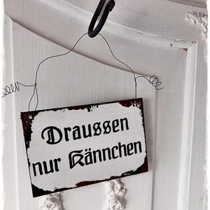 Könnte beinhalten: Ein weißes Metallschild mit schwarzem Text, der "Draussen nur Känmchen" lautet. Das Schild hängt an einem schwarzen Haken an einer weißen Tür.