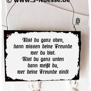 May include: A rectangular sign with a black border and white interior, featuring German text. The sign is suspended by a wire. The text reads, "If you're at the top, your friends know who you are. If you're at the bottom, you know who your friends are!"