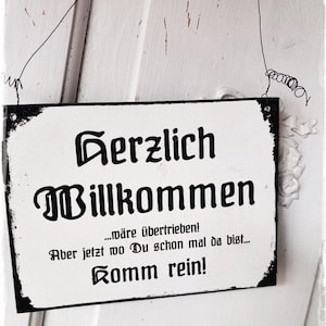 以下が含まれることがあります： 「Herzlich Willkommen...wäre übertrieben! Aber jetzt wo Du schon mal da bist...Komm rein! 」と黒文字で書かれた白い看板と、著作権記号と「3NUESSE」の文字。