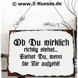 May include: A white sign with black text that reads "Ob Du wirklich richtig stehst... Siehst Du, wenn die Tür aufgeht!" The sign is hanging from a wire and has a rustic, distressed look.