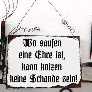 May include: A white metal sign with black text that reads "To saufen eine Ehre ist, kann kotzen keine Schande sein!" The sign is hanging from a wire.