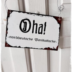 以下が含まれることがあります： 「Oh! -norddeutsche Panikattacke-」と黒文字で書かれた白い金属製の看板。看板は古びてさび付いたような外観で、ワイヤーで吊り下げられている。