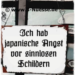May include: A white sign with black text that reads "Ich hab japanische Angst vor sinnlosen Schildern". The sign is attached to a wooden surface with a rusty metal hinge.