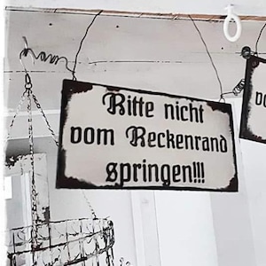 Può includere: Un cartello bianco e nero con il testo "Bitte nicht vom Reckenrand springen!!!" appeso a un soffitto bianco.