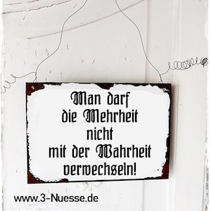 Könnte beinhalten: Ein rechteckiges Schild mit weißem Hintergrund und dunkelrotem Rand. Das Schild trägt schwarzen Text mit der Aufschrift "Man darf die Mehrheit nicht mit der Wahrheit verwechseln!" und "www.3-Nuesse.de". Das Schild wird von einem Draht gehalten.
