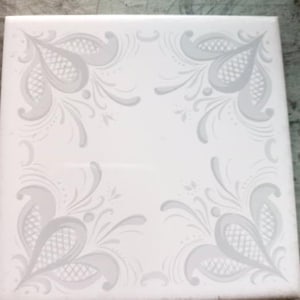 Puede incluir: Azulejo blanco cuadrado con un diseño floral gris decorativo en cada esquina. El diseño presenta hojas estilizadas y formas de tipo paisley. El azulejo es probablemente para usar en una cocina o baño.