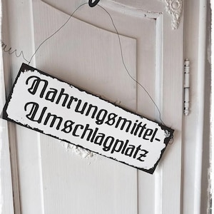 以下が含まれることがあります： 白いドアに黒い文字の白い看板が掛かっています。看板には「Nahrungsmittel- Umschlagplatz」と書かれています。
