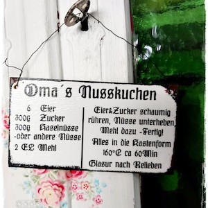 Könnte beinhalten: Ein weißes Schild mit schwarzem Text, auf dem "Oma's Nusskuchen" steht und die Zutaten und Anweisungen für die Herstellung eines Nuss Kuchens aufgelistet sind. Das Schild hängt an einer weißen Tür mit einem Schlüsselloch.