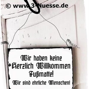 May include: A black and white sign with the text "Wir haben keine Herzlich Willkommen Fußmatte! Wir sind ehrliche Menschen!" hanging on a white door.