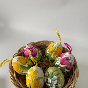 Könnte beinhalten: Ein Weidenkorb enthält sechs handbemalte Ostereier. Die Eier weisen florale Muster in Gelb, Rosa und Grün auf. Jedes Ei hat eine Schleife zum Aufhängen. Der Korb ist aus hellbraunem Korbgeflecht.